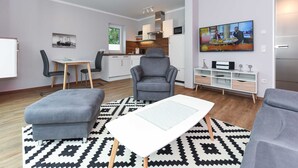 Een moderne woonkamer met een grijze bank, een voetenbankje en een witte salontafel. Er is een keuken met witte kastjes en een eettafel met stoelen. Een flatscreen televisie hangt aan de muur, en er is een schilderij te zien.