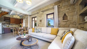 A modern living room with a stone wall, a sofa, a dining area with a table and chairs, and a kitchen in the background. - Asion Villa Orion with Private Pool (Kato Asites)