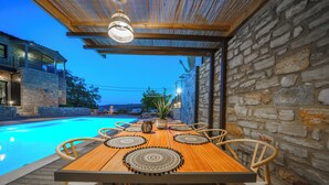 A wooden poolside table with woven placemats, surrounded by wicker chairs, set against a stone wall and under a wooden pergola with a hanging light fixture. - Asion Villa Orion with Private Pool (Kato Asites)
