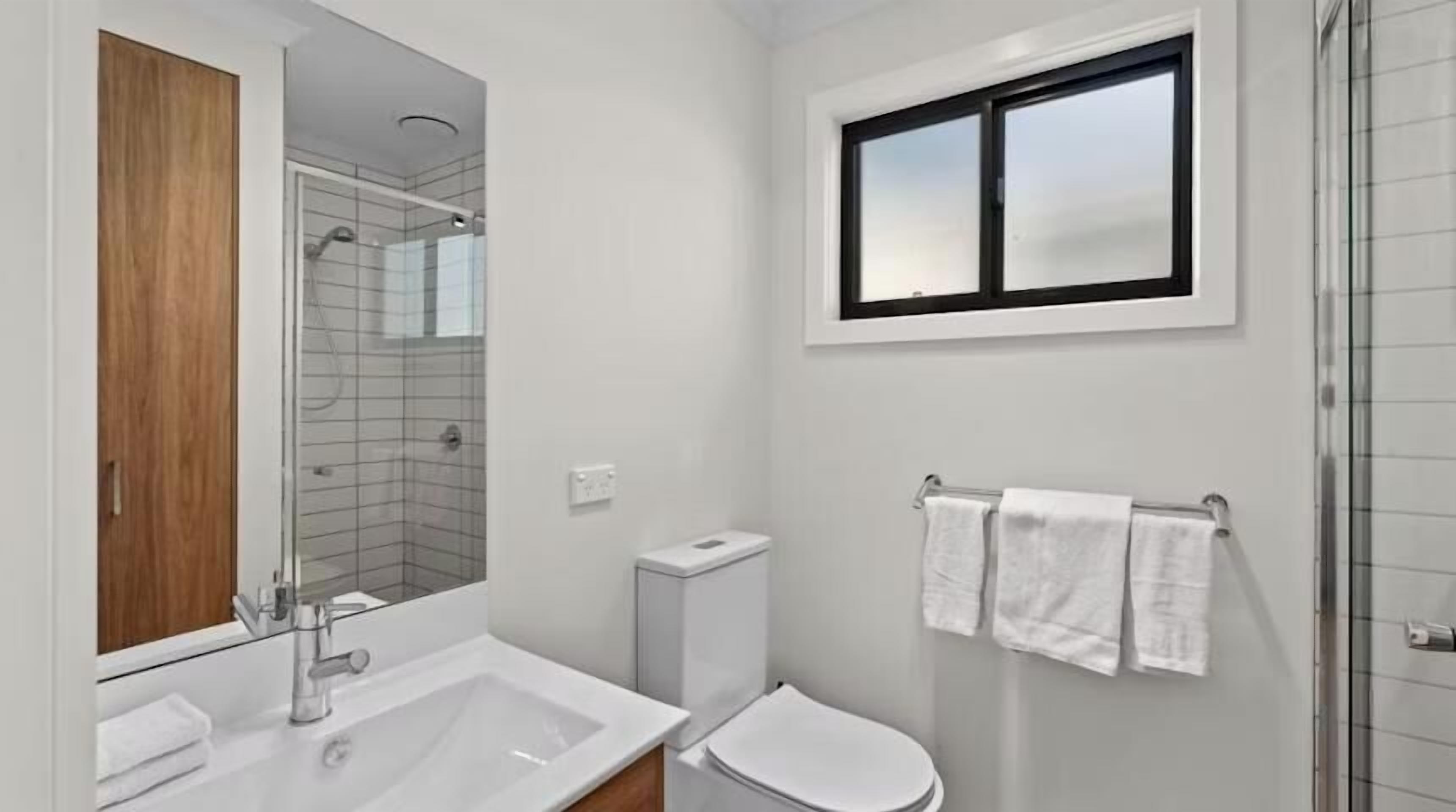 A modern bathroom with a white sink, mirror, and toilet. There is a window above the toilet and a shower area with glass doors.
