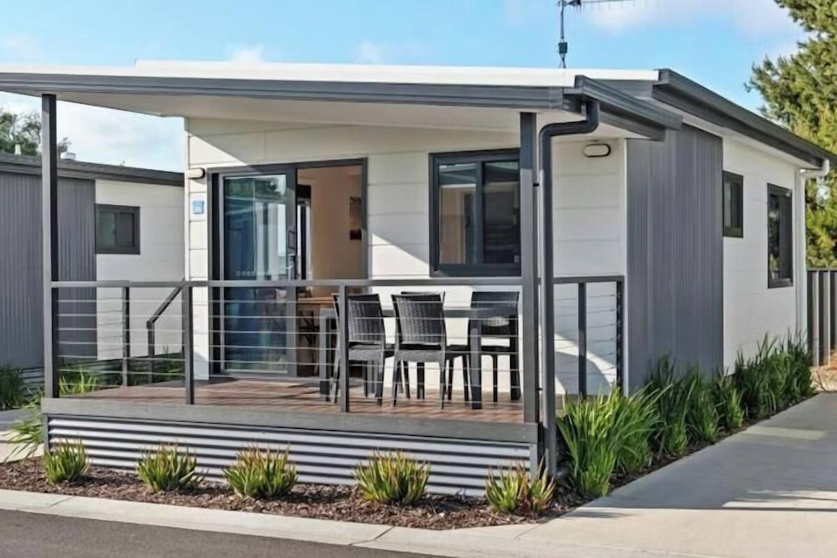 A modern, single-story mobile home with a covered patio area, a small outdoor seating arrangement, and a visible entrance.