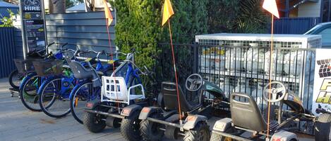 A row of ATVs and bicycles parked in a lot with a signboard and a fence in the background.