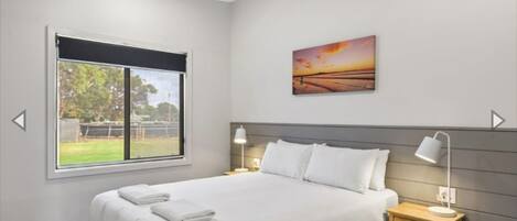 A bedroom with a large bed, a nightstand with a lamp, a window with a view of greenery, and a sunset painting on the wall.