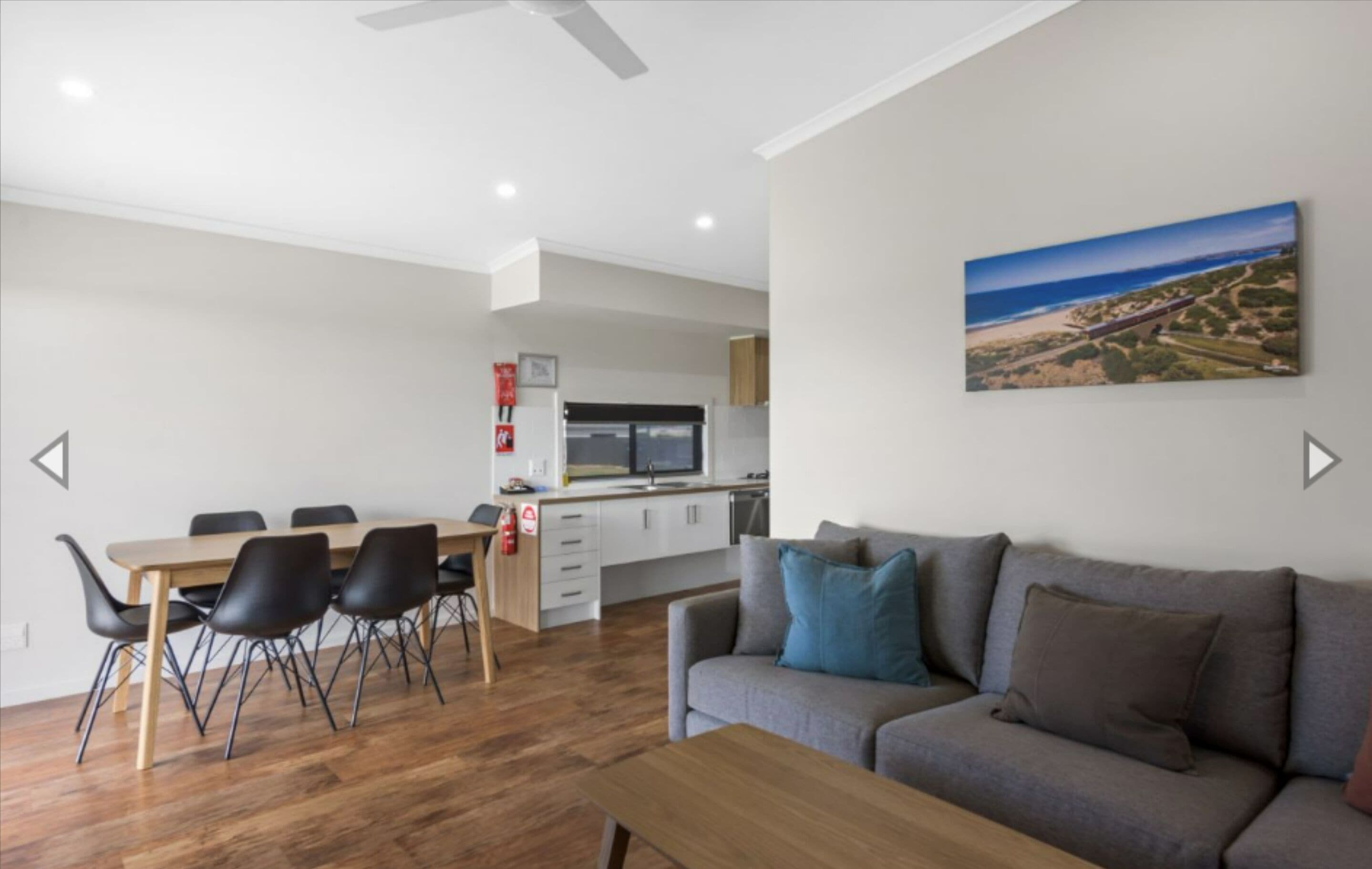 Una sala de estar moderna con un sofá gris, una mesa de centro de madera, y un comedor con una mesa de madera y sillas negras. Se ve una cocina con armarios blancos y un microondas al fondo.