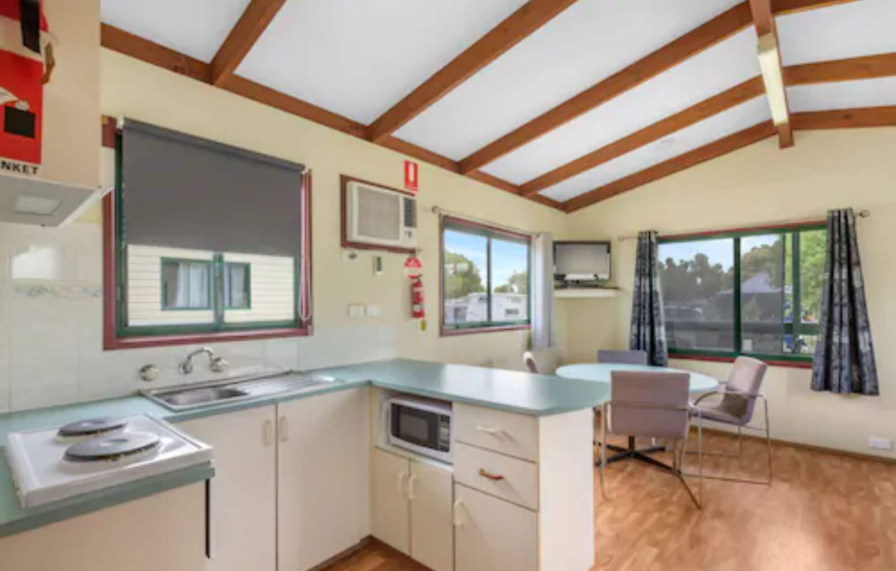 A compact kitchen with a microwave, sink, and cabinets, a dining area with a table and chairs, and a large window with curtains.