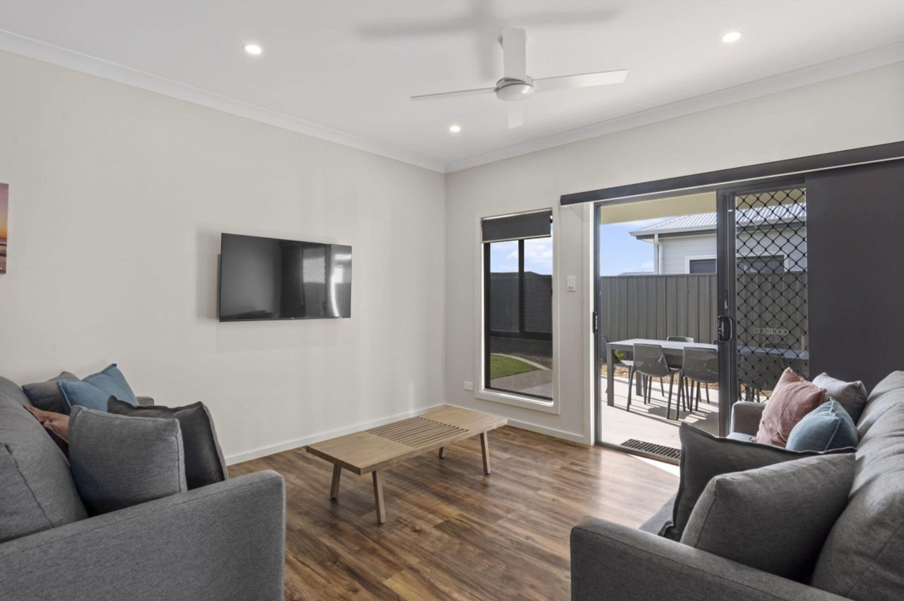 A modern living room with a flat-screen TV, grey sofas, a wooden coffee table, and a sliding glass door leading to an outdoor patio.