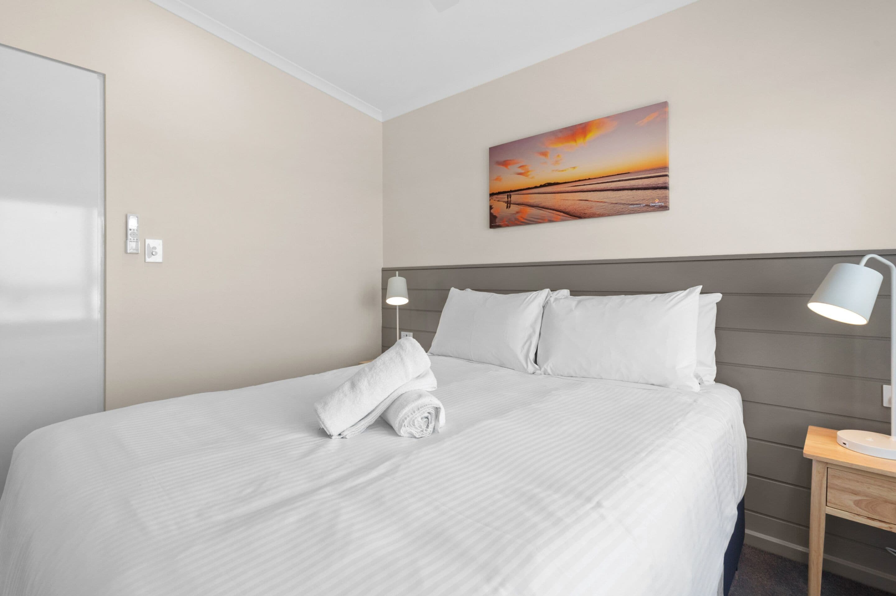 Una cama bien hecha con sábanas y almohadas blancas, una lámpara de noche y un cuadro de un atardecer en la pared.