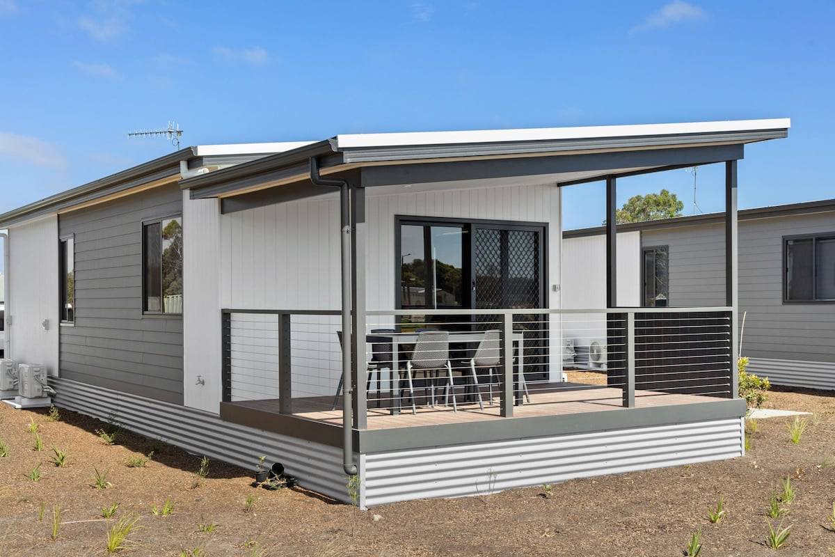 A modern single-story mobile home with a covered patio area, a small outdoor kitchen, and a fenced deck.