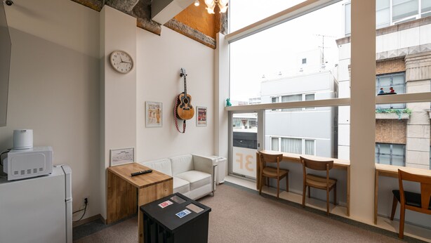 Una habitación moderna con un sofá blanco, sillas de madera, una pequeña mesa, una guitarra y un gran ventanal con vistas a edificios.