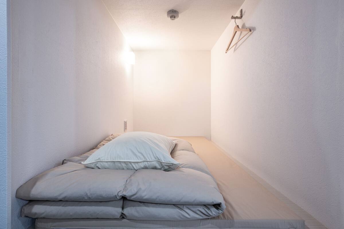 A narrow, well-lit room with a bed at the end, a light fixture on the ceiling, and a wall-mounted hook.