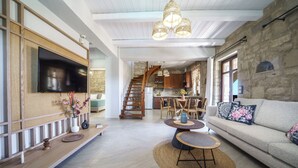 Un vaste espace de vie comprenant un canapé, une table basse et un téléviseur. Un escalier mène à une cuisine dotée de meubles en bois et d’un mur en pierre.
