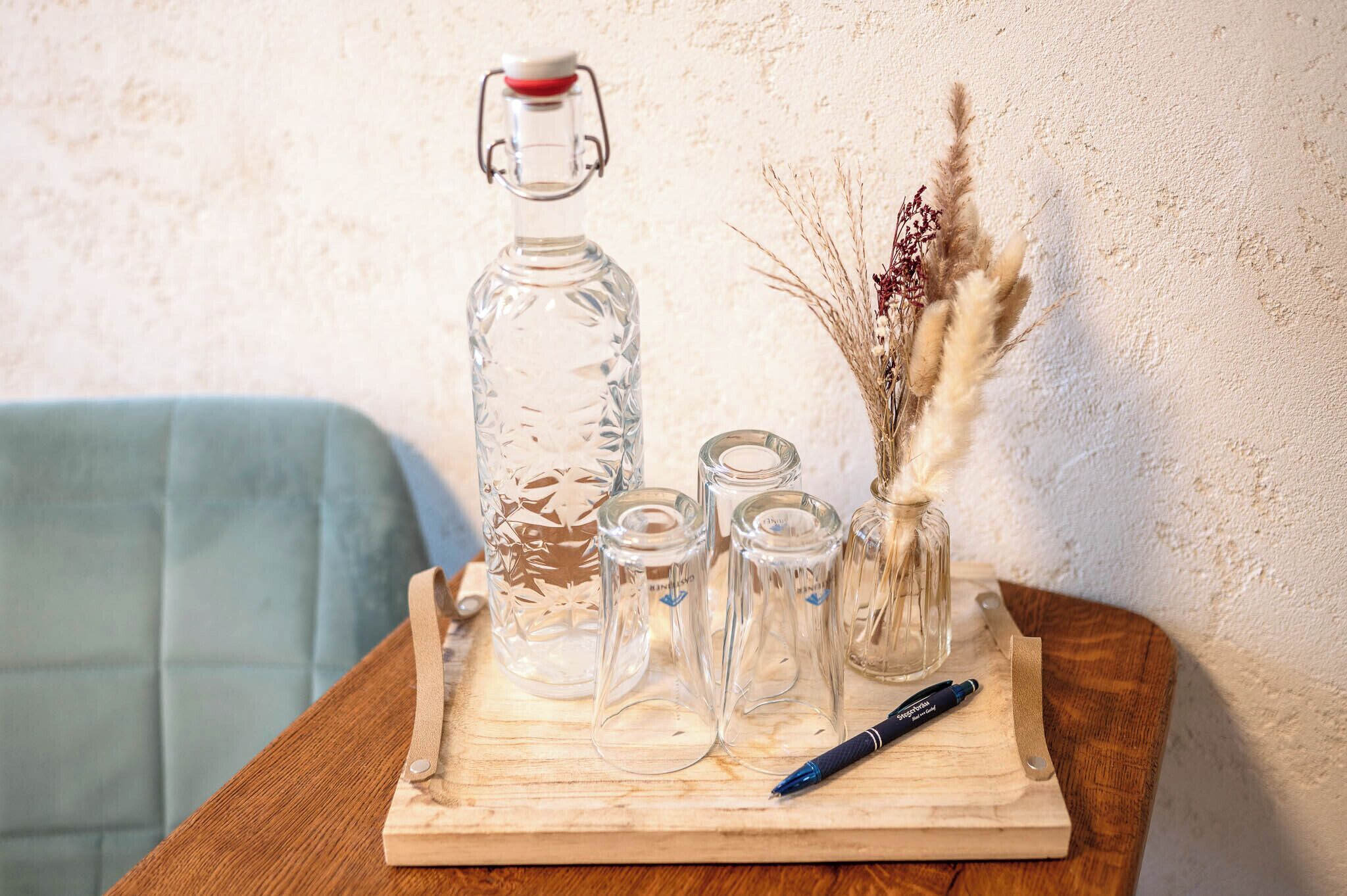 A wooden tray with a glass decanter, small glass bottles, a pen, and dried plants on a table.