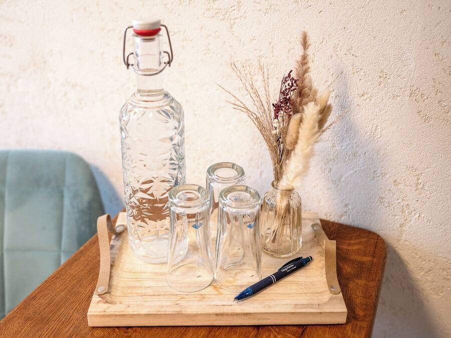 A wooden tray with a glass decanter, small glass bottles, a pen, and dried plants on a table.