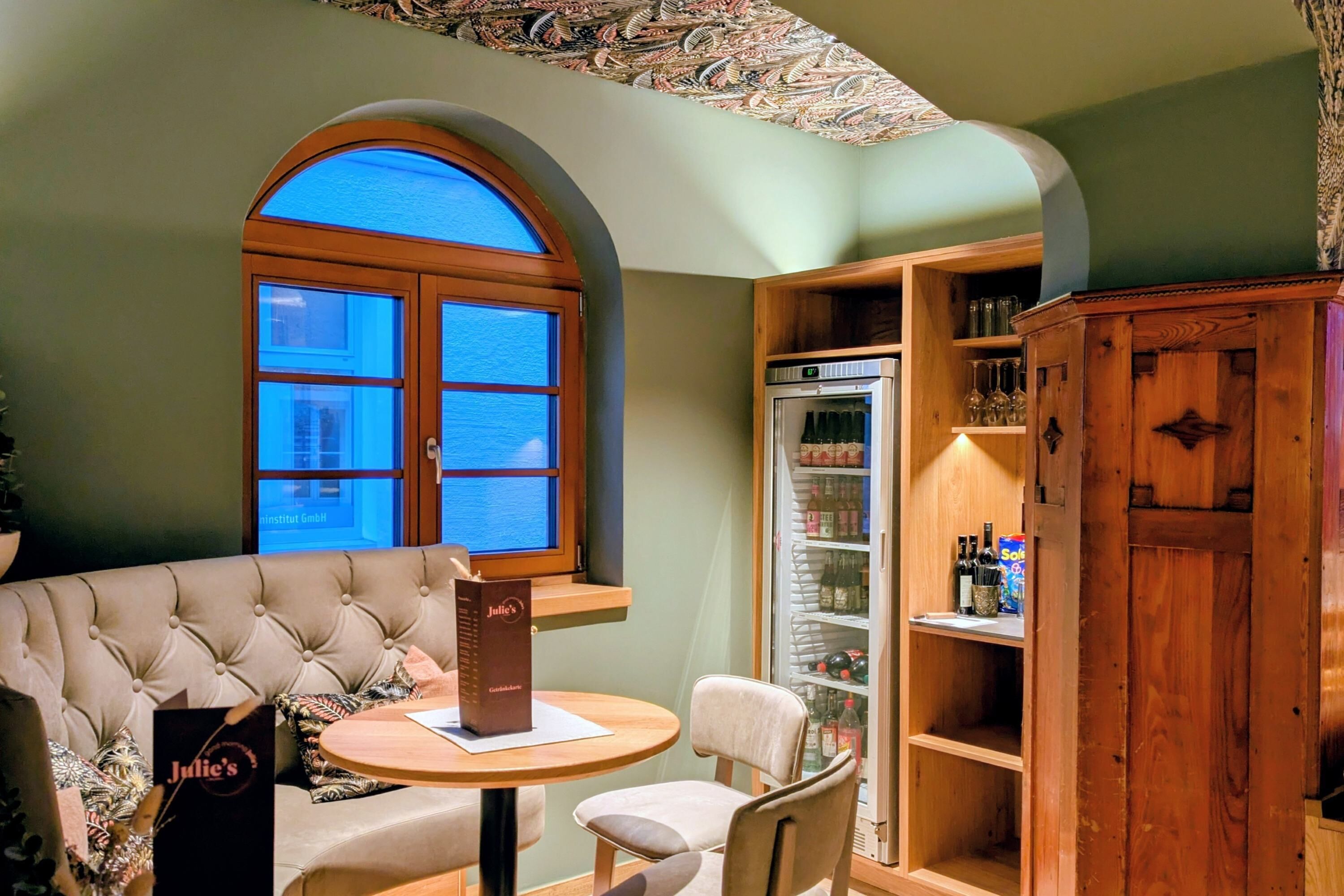A corner room with a window, a refrigerator, a wooden cabinet, and a table with a chair.