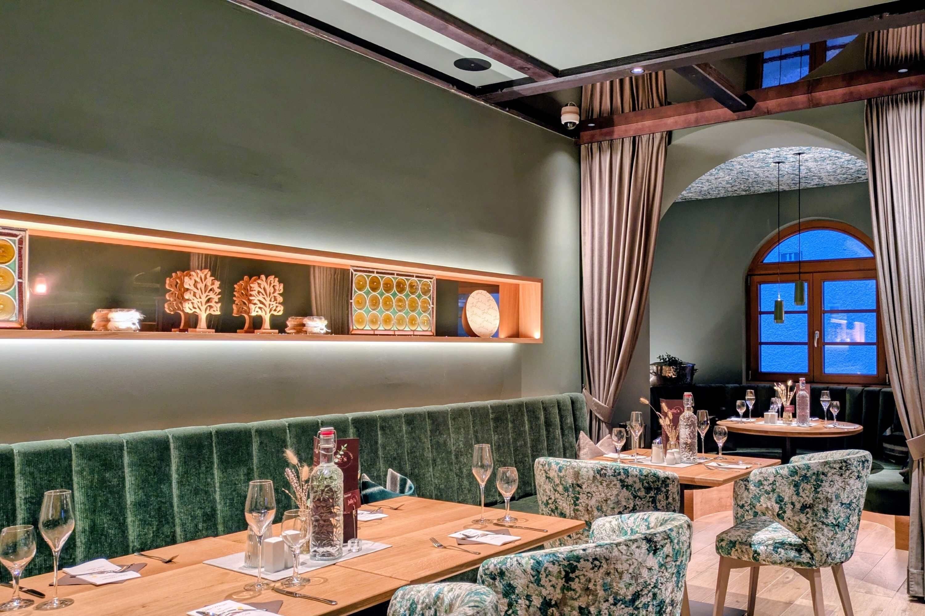A restaurant with a green upholstered bench seating area, wooden tables, and floral-patterned chairs.
