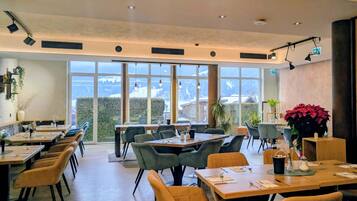 A modern dining area with tables and chairs, large windows, and indoor plants.