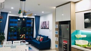 A modern living room with a blue sofa, a dining table with chairs, a kitchen with a refrigerator, and a window with blue curtains. - Park Hill Times City Apartment						 						 (Hanoi)