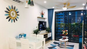 A modern living room with a glass dining table, white chairs, a TV, and a decorative sun-shaped clock. - Park Hill Times City Apartment						 						 (Hanoi)