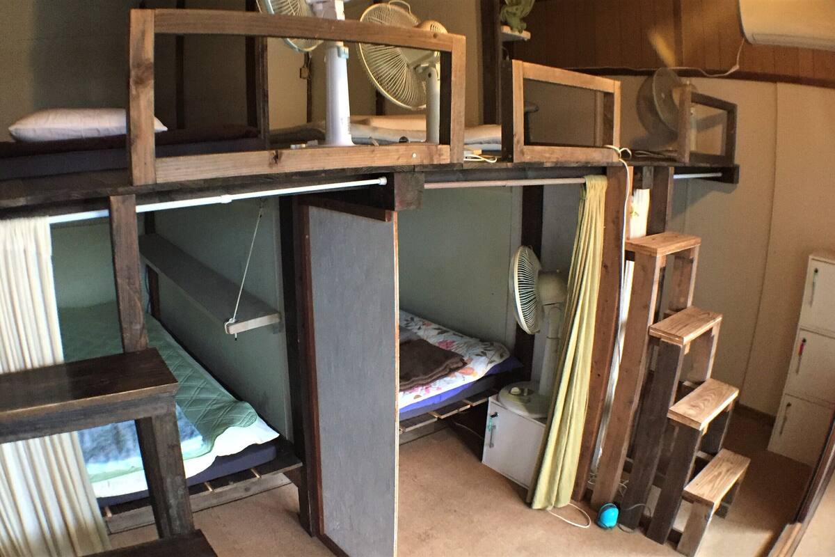 Una habitación pequeña con literas, un escritorio, una silla, un ventilador y una ventana.