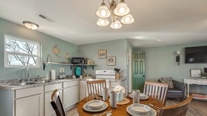 Una cocina con armarios blancos, una mesa de comedor con sillas, una araña y un televisor al fondo.