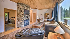 A cozy living room with a stone fireplace, leather sofa, and a large window offering a view of the forest. - Secluded Camp Connell Home w/ Deck + Fireplace! (Camp Connell)