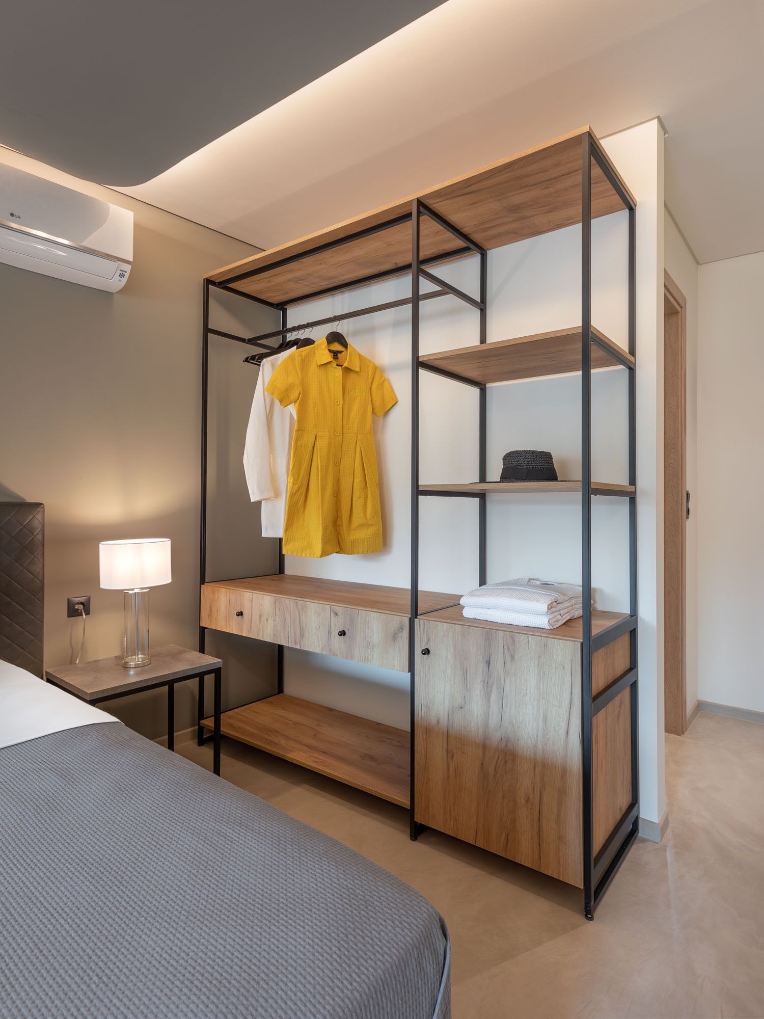 A modern bedroom with a bed, a bedside table with a lamp, a wooden wardrobe with shelves, and a hanging rack with clothes.
