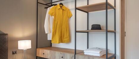 A modern bedroom with a bed, a bedside table with a lamp, a wooden wardrobe with shelves, and a hanging rack with clothes.