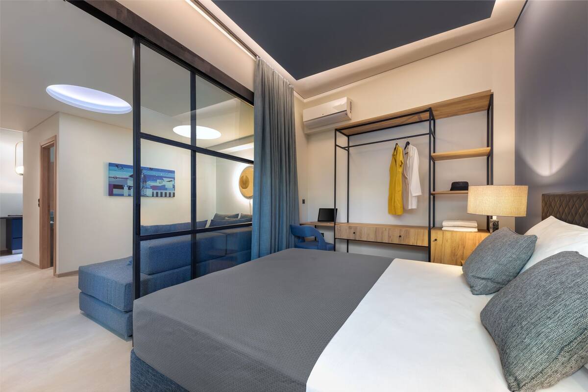 Une chambre d’hôtel moderne dotée d’un grand lit, d’un bureau, d’une chaise, d’une armoire et d’une salle de bain visible à travers une paroi en verre.