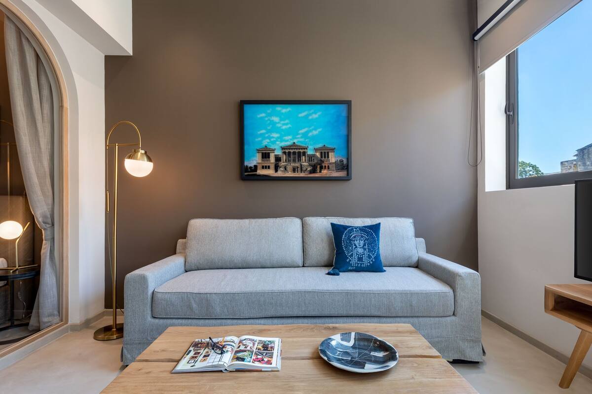 Un salon moderne avec un canapé gris, une table basse sur laquelle reposent un magazine et une assiette décorative, un lampadaire et un tableau encadré accroché au mur.