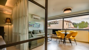 Un salon moderne avec un canapé, une table à manger entourée de chaises jaunes et une grande fenêtre donnant sur des immeubles et des arbres.