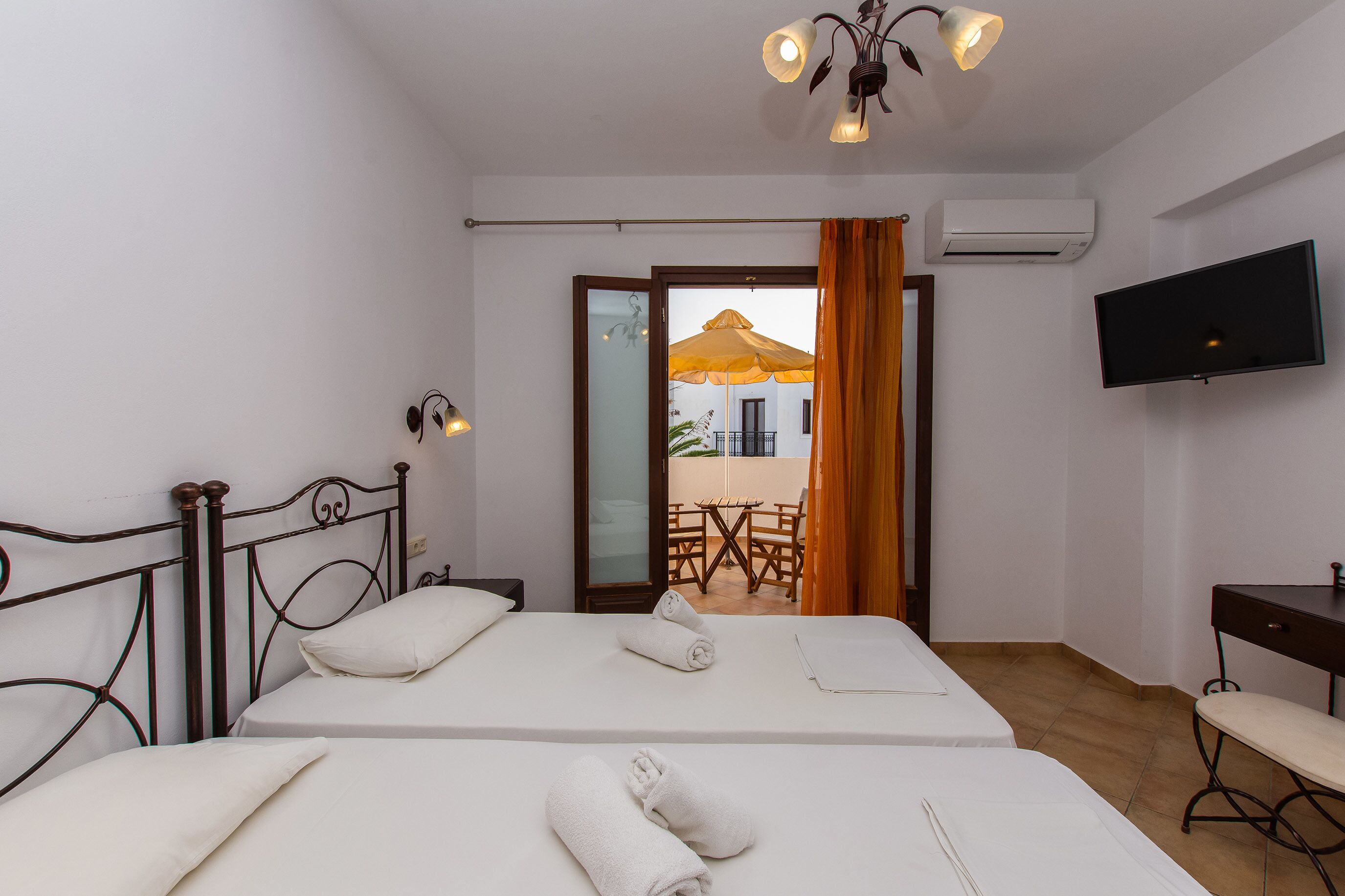 Ein Hotelzimmer mit zwei Betten, einem Fernseher, einem Schreibtisch und einem Balkon mit Sonnenschirm.
