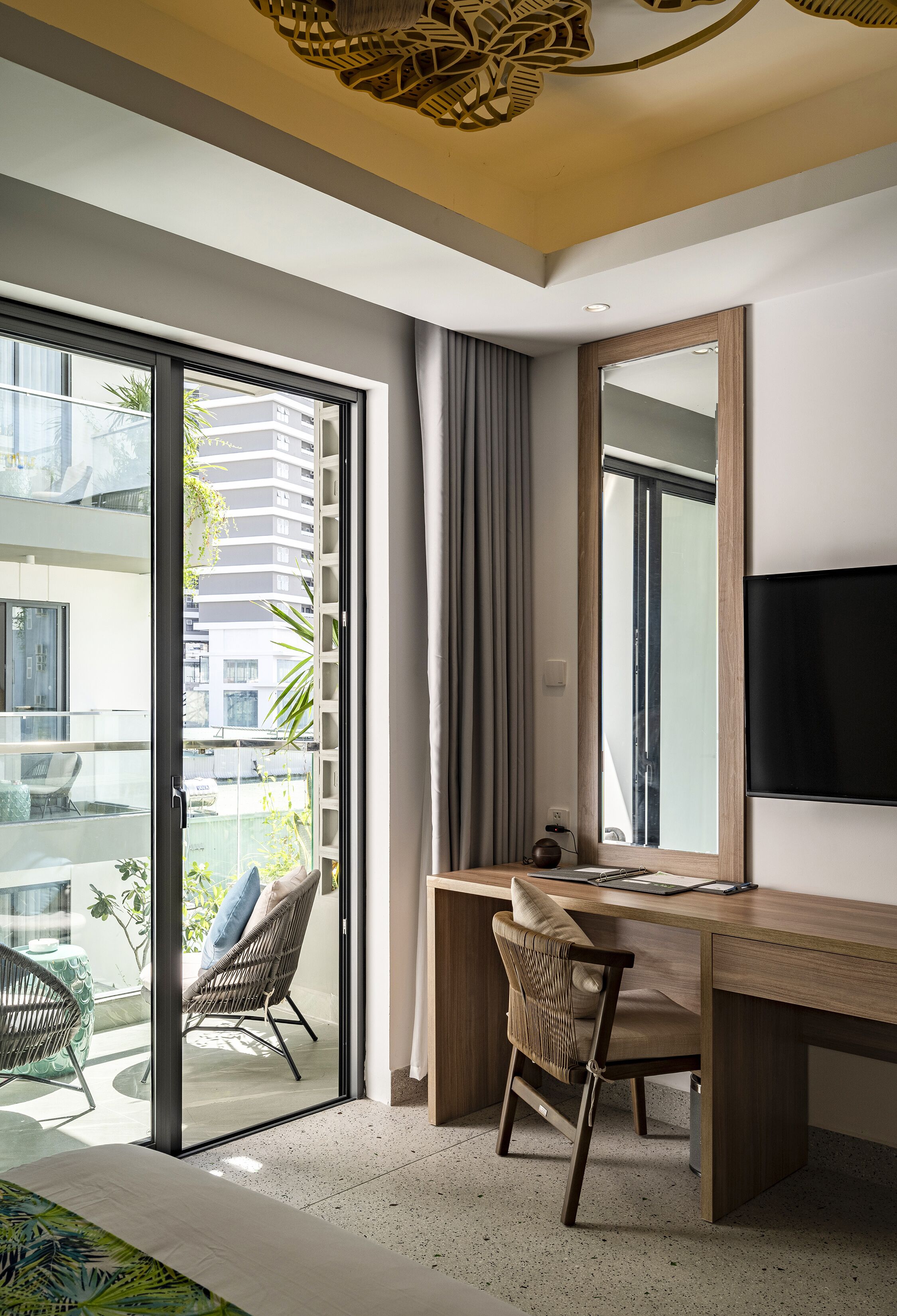 A modern hotel room with a large sliding glass door leading to a balcony, a wooden desk, two wicker chairs, a flat-screen TV mounted on the wall, and a ceiling light fixture.