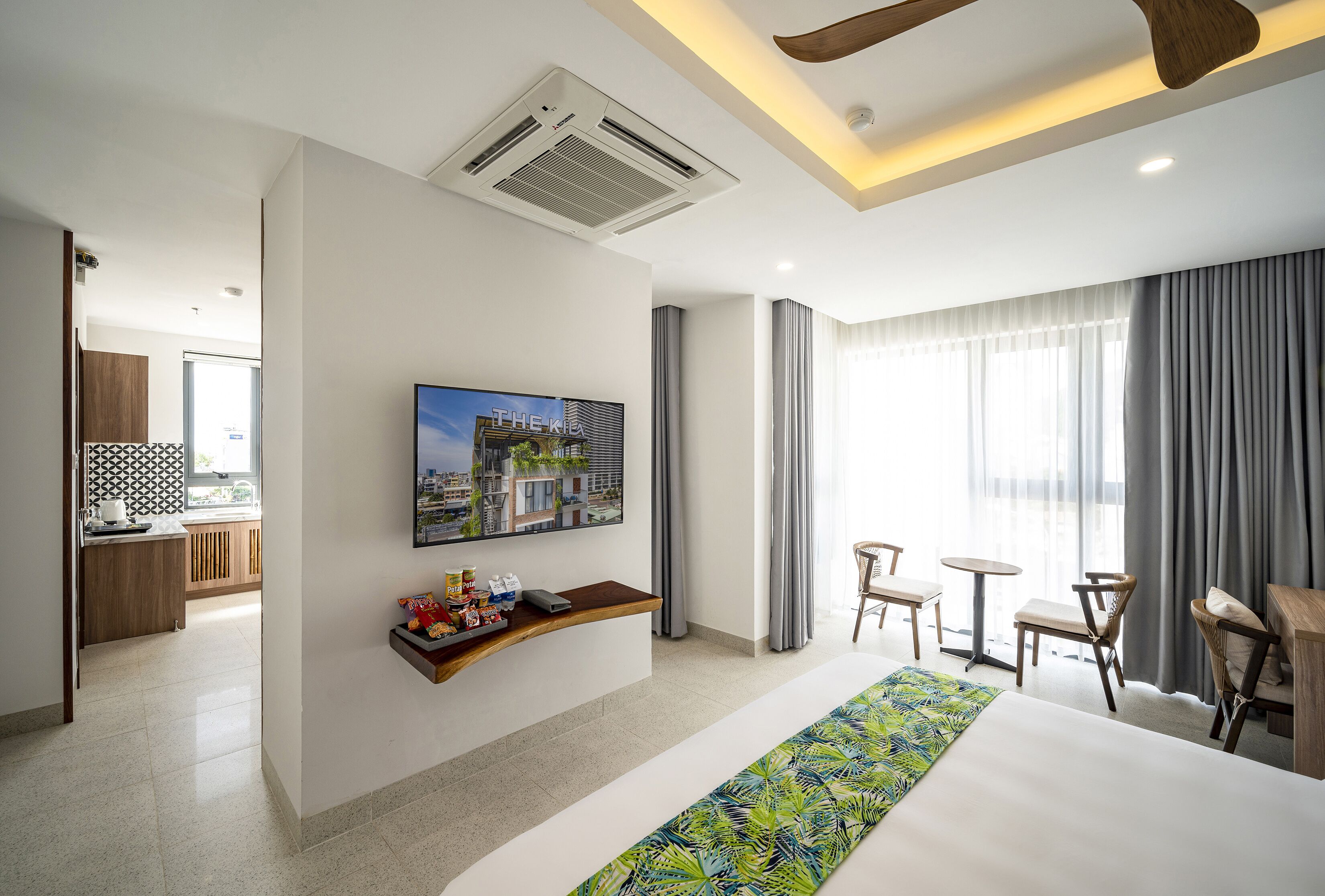 A hotel room with a large bed, a TV mounted on the wall, a small kitchen area, and a balcony with curtains.