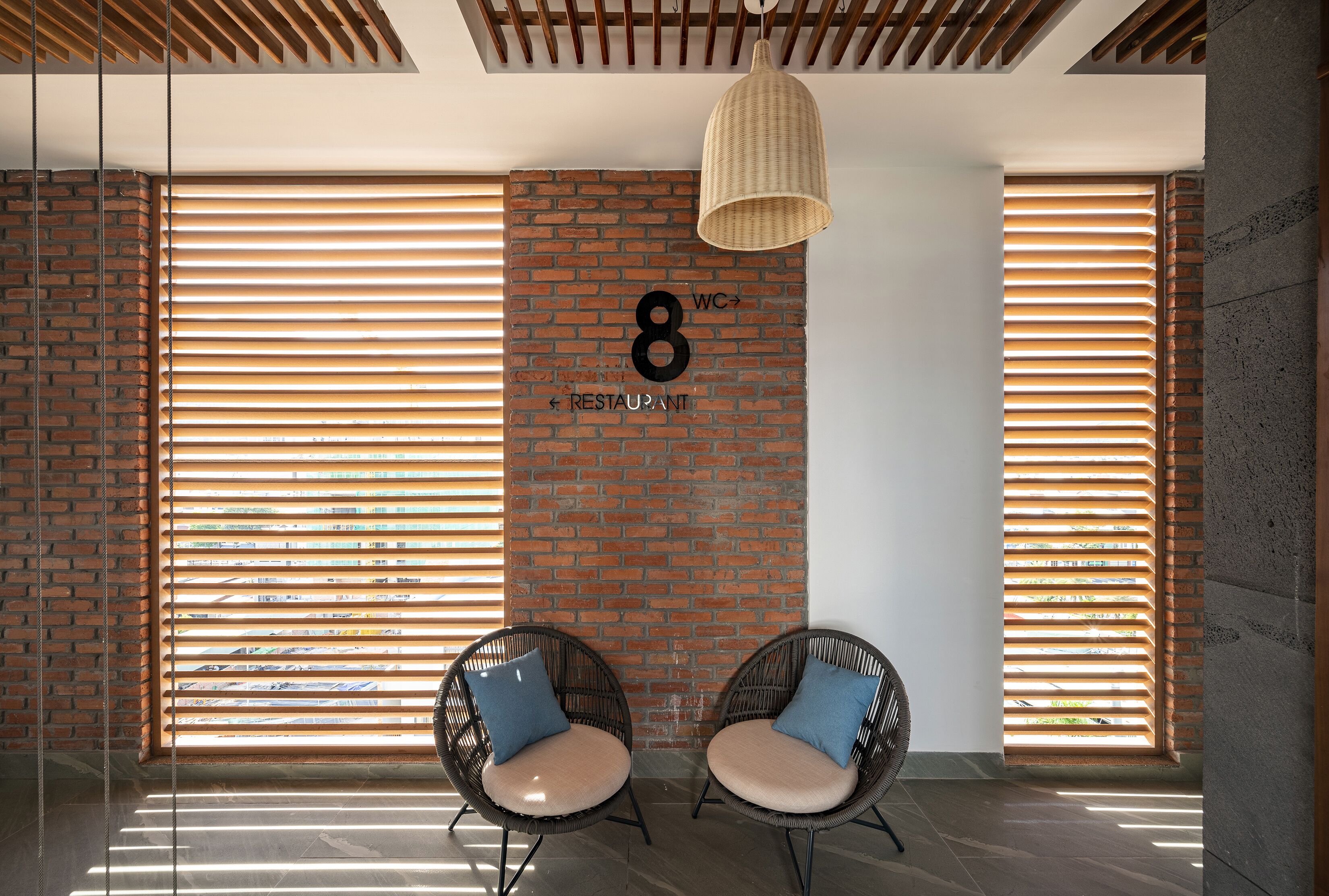 A modern interior with a brick wall, two wicker chairs with cushions, a hanging light fixture, and a sign indicating the location of the restroom and restaurant.
