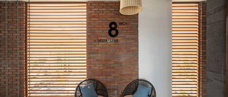 A modern interior with a brick wall, two wicker chairs with cushions, a hanging light fixture, and a sign indicating the location of the restroom and restaurant.