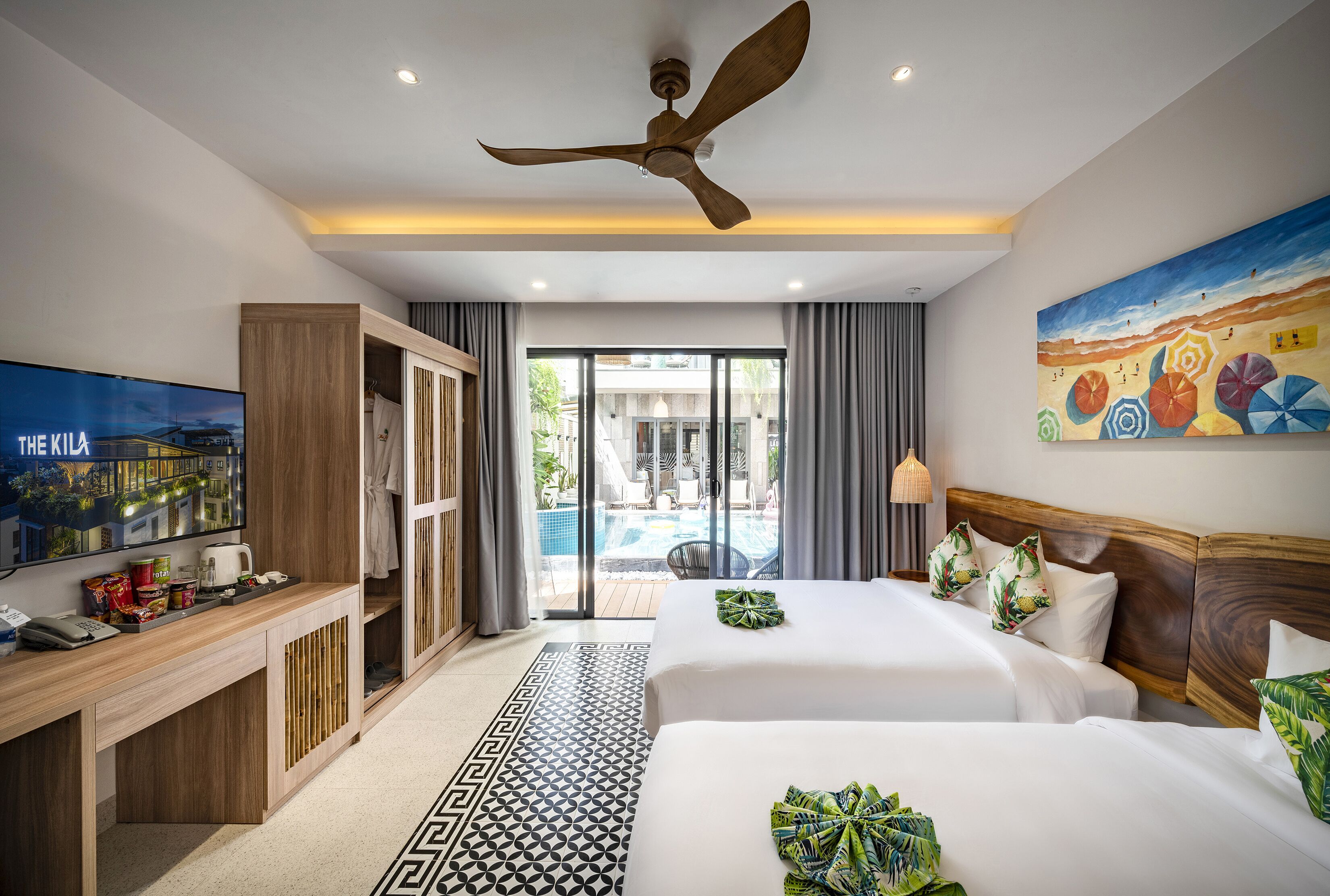 A hotel room with two beds, a TV, a desk, a ceiling fan, and a view of the pool.