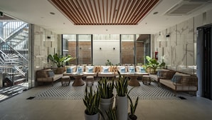 Een moderne lobby met een houten plafond, grote ramen en een zithoek met rieten meubels en plantenpotten.