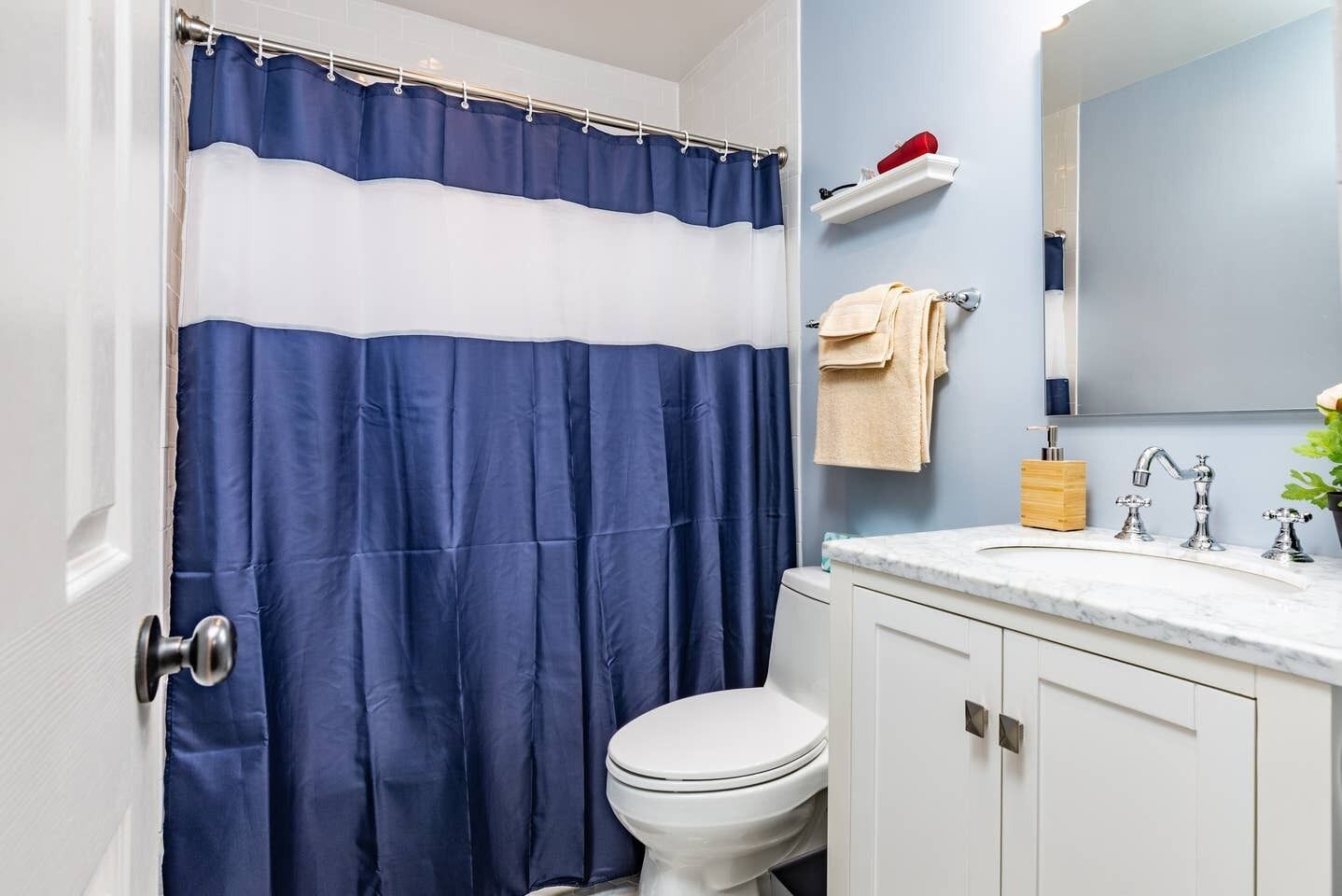 A bathroom with a blue and white shower curtain, a white toilet, a marble sink with two faucets, a wooden soap holder, and a towel rack with a beige towel.