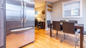 A modern kitchen with a stainless steel refrigerator, wooden flooring, a dining area with a table and chairs, and a window with blinds.