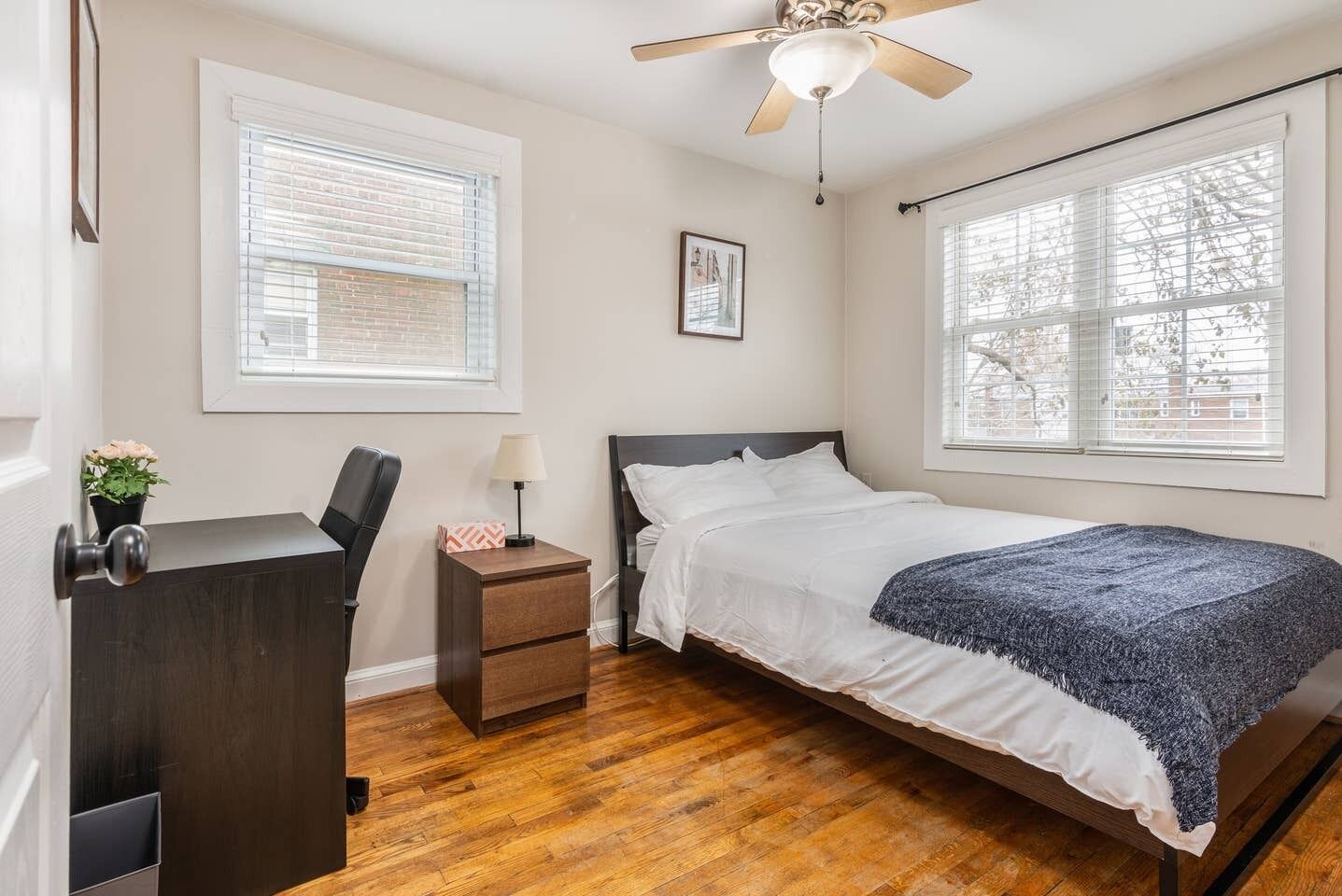 A bedroom with a bed, a desk, a chair, a nightstand, a ceiling fan, and two windows with blinds.