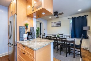 A modern kitchen with wooden cabinets, a granite countertop, and a coffee machine. There is a dining area with a table and chairs, and a ceiling fan above.