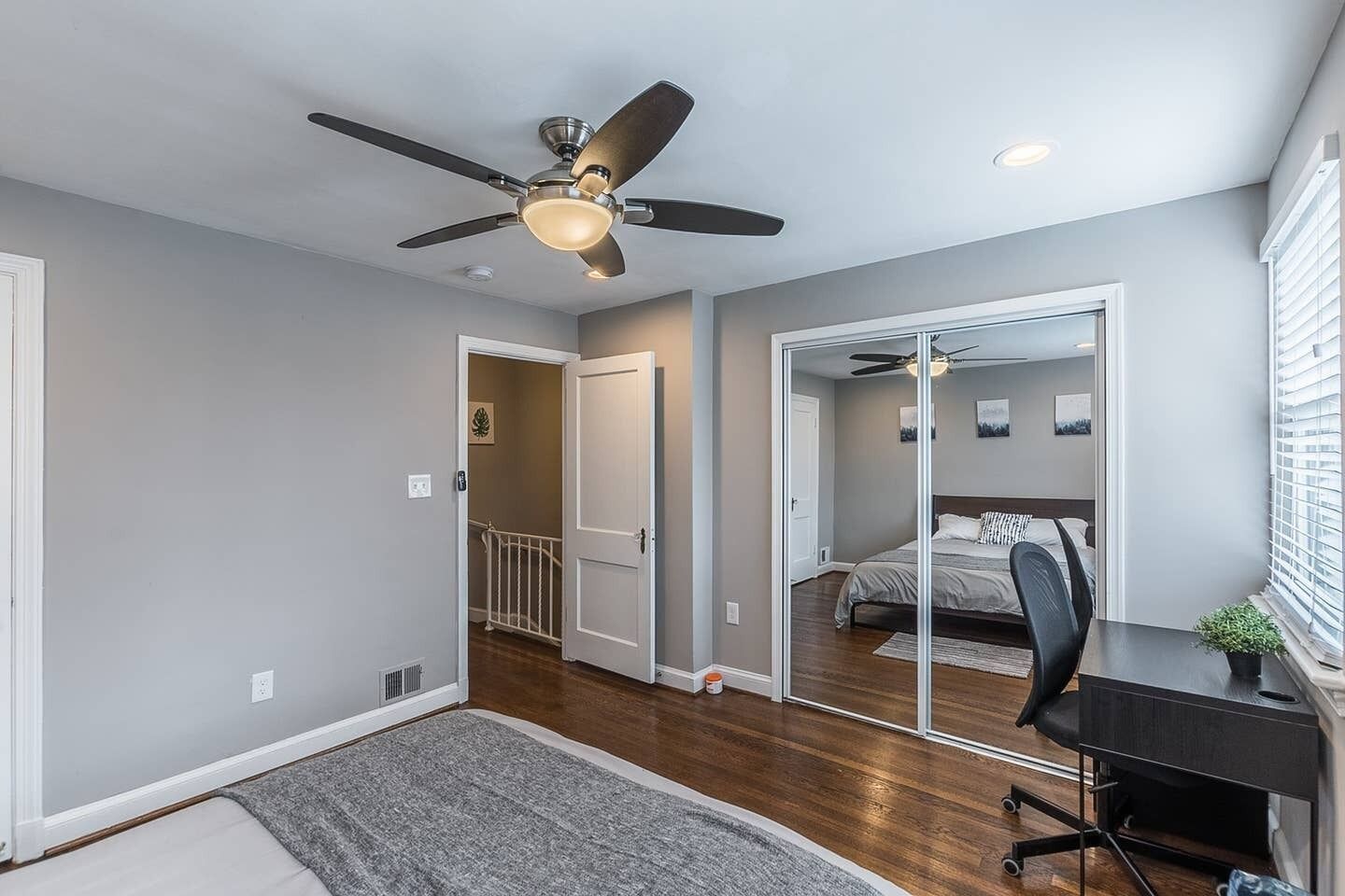 A bedroom with a bed, a desk, and a chair. There is a ceiling fan and a window with blinds.