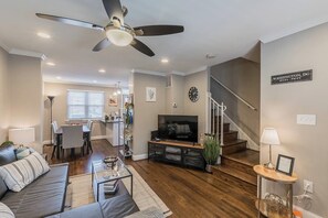 A modern living room with a sofa, coffee table, TV, and a staircase leading to another level. - Beautiful 3br/2ba House with cozy backyard by CozySuites (Alexandria)
