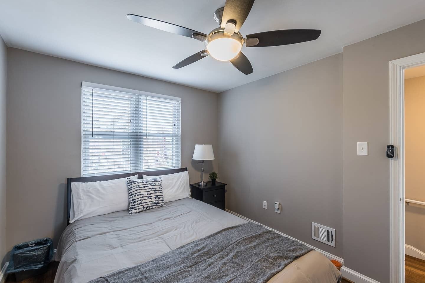 A bedroom with a bed, a ceiling fan, a window with blinds, a nightstand with a lamp, and a door.