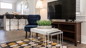 A modern living room with a dark blue armchair, a white coffee table with a plant, a large flat-screen TV, and a patterned rug. - Louisville Dream House 4BR/2BA  by CozySuites (Louisville)