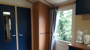 Een kamer met een houten garderobe, een spiegel, een raam met gordijnen, een koffiezetapparaat en een radiator.