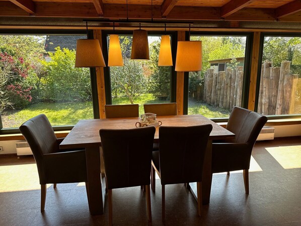 A dining area with a wooden table and four chairs, large windows with curtains, and hanging lamps.