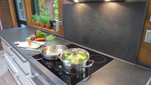 Eine Küche mit einem schwarzen Induktionskochfeld, einem Topf mit Deckel, einem Schneidebrett mit Gemüse und einem kleinen Fenster mit Blumentöpfen.