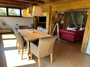 A wooden dining table with chairs, a red sofa, and a lamp in a room with wooden beams and a stone wall.