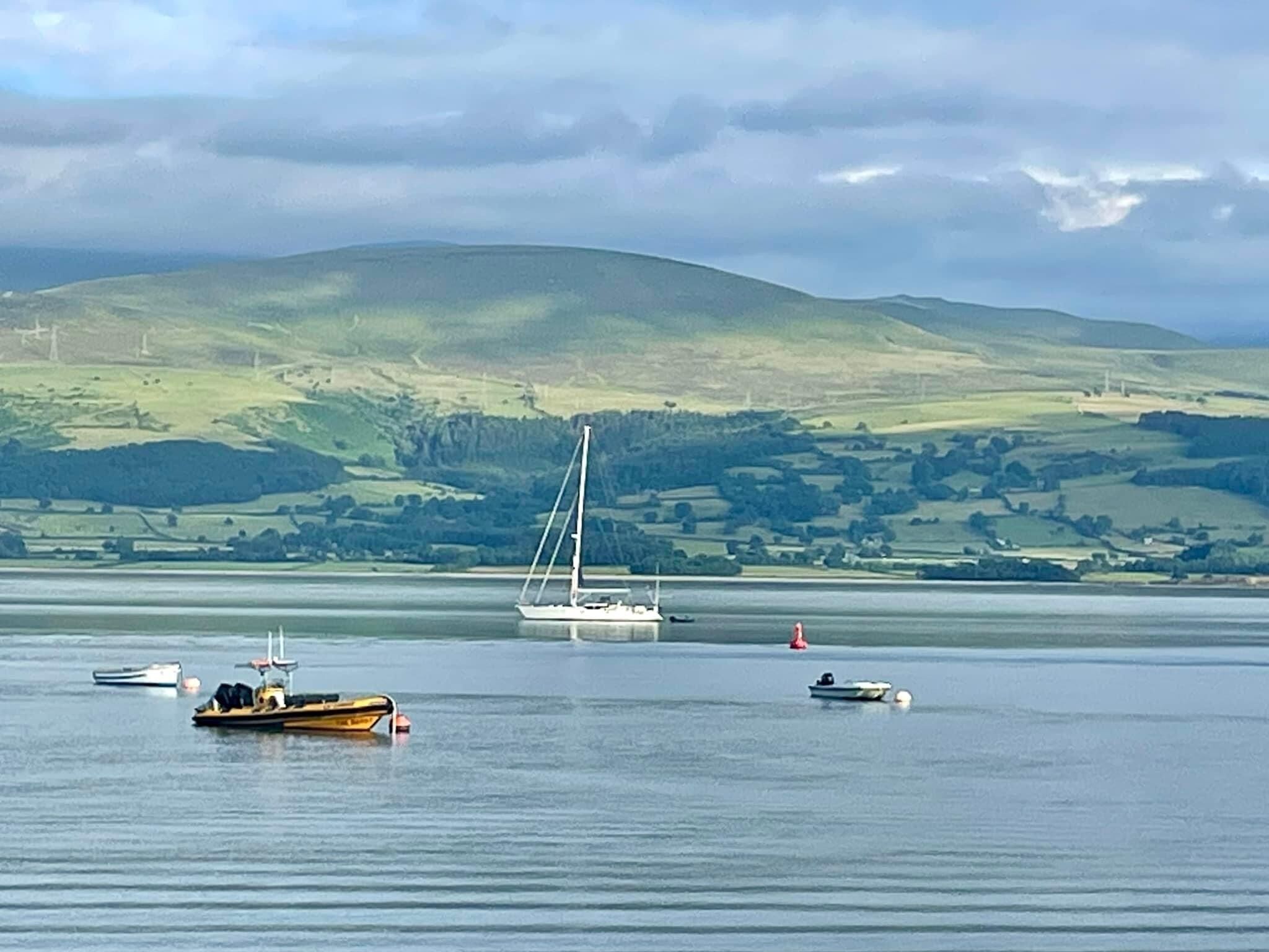 Un lac paisible où flottent plusieurs bateaux, dont un voilier et un bateau à moteur, et où l'on aperçoit au loin des collines ondoyantes.